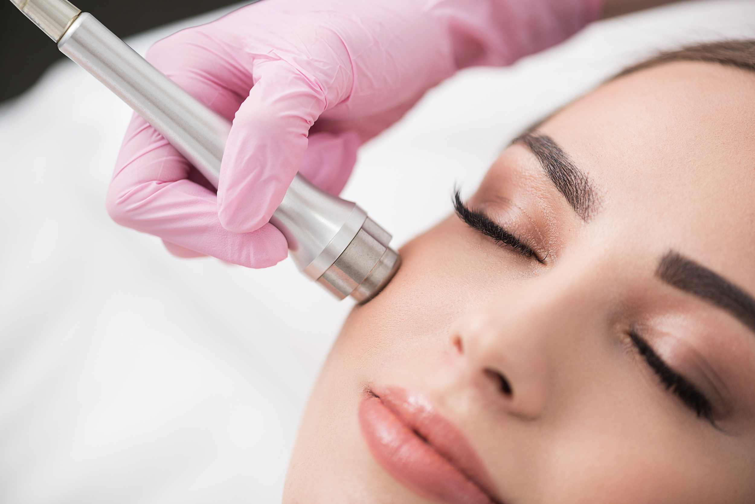 Close up of instrument in hand of doctor-cosmetologist, which is making procedure microdermabrasion of facial skin for beautiful young woman at beauty salon. Girl is lying with closed eyes